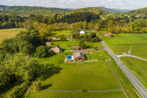 Aerial view of a stone farmhouse and red barn along a country road framed by mountains and trees