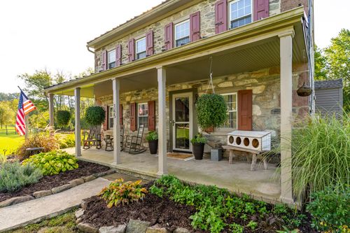 Covered porch of a stone farmhouse with red shutters, rocking chairs and hanging planters