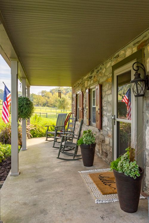 Covered stone farmhouse porch with rocking chairs, wind chimes and a view of fenced pasture