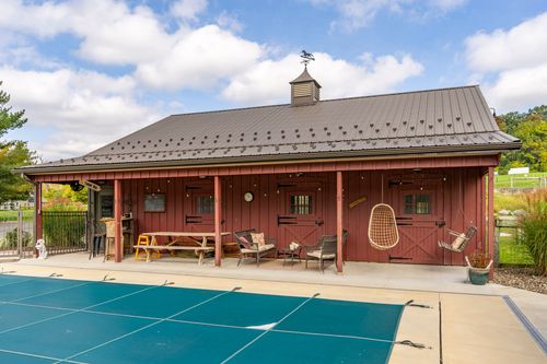 Red barn with a covered porch, picnic table seating and rocking chairs beside a pool