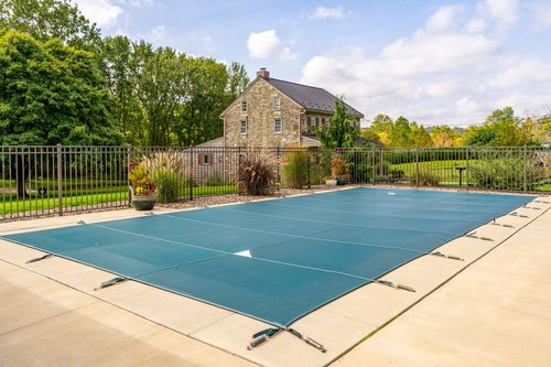 Covered pool with a stone farmhouse visible beyond a wrought iron fence and landscaping