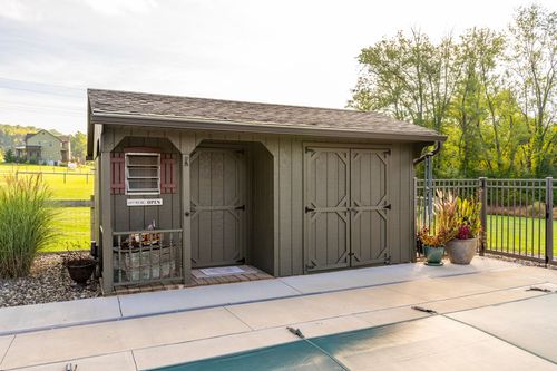 Gray storage shed with double doors and a shingled roof beside a covered pool