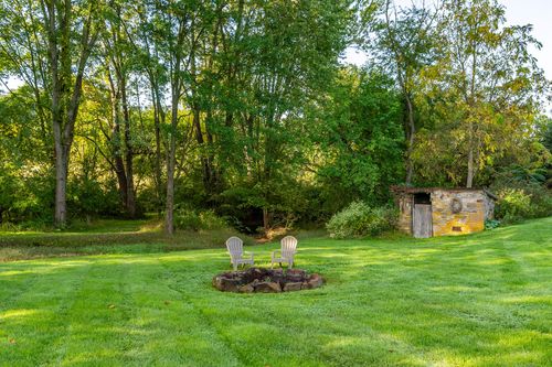 Fire pit with wooden chairs set in an open lawn near a stone outbuilding and trees