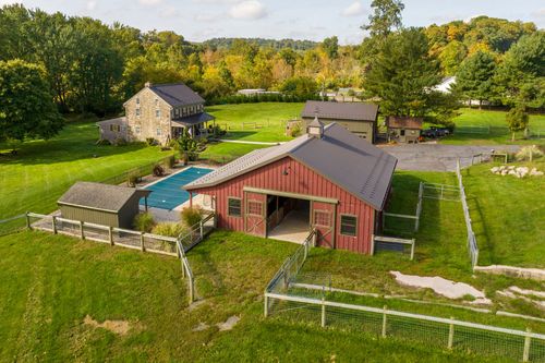 Aerial view of a red bank barn beside a stone farmhouse with a pool and detached garage