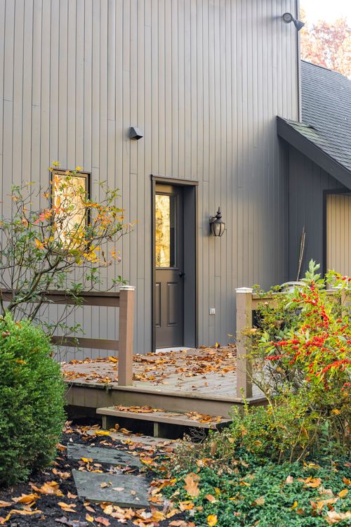 Rear deck entry with gray door, sconce light and railing among autumn shrubs