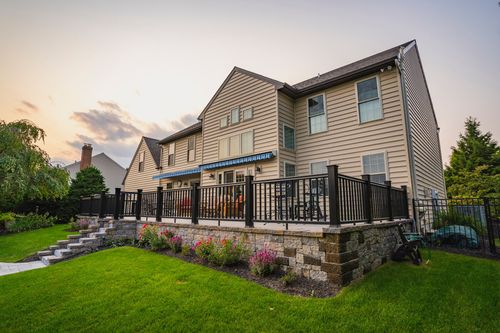 Two-story tan home exterior with black metal deck railing and stone retaining wall