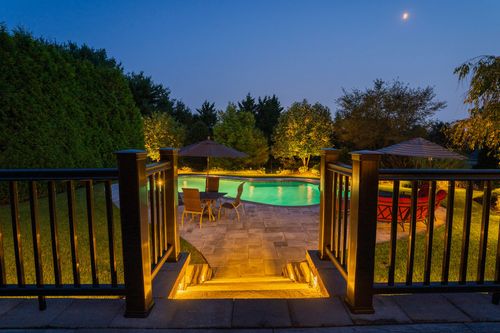 Backyard deck steps down to an illuminated pool patio with red seating at night