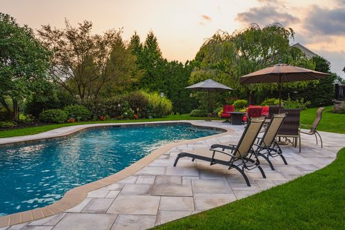 Backyard pool with stone patio, lounge chairs and umbrellas at dusk with home in view