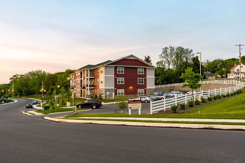 Three-story apartment building along a suburban road at dusk with a white picket fence and wooded backdrop
