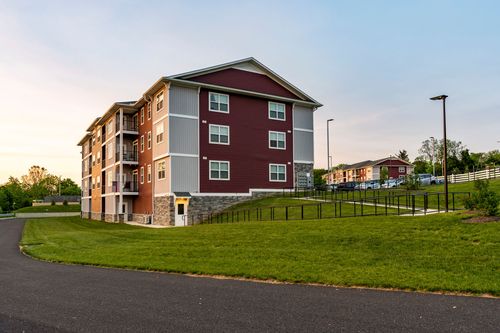 Corner elevation of a red apartment building on a grassy slope framed by a black metal fence at dusk