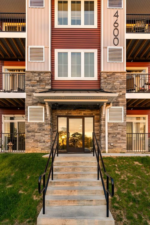 Apartment building entrance with glass doors reflecting a sunset sky, flanked by stone columns and twin stair railings
