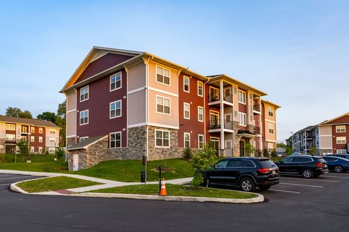 Apartment building corner on a grassy lot with a curving walkway and cars parked at dusk