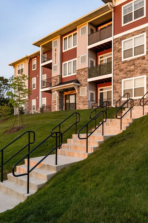 Apartment building corner with balconies and a black-railed staircase climbing a landscaped hill at dusk