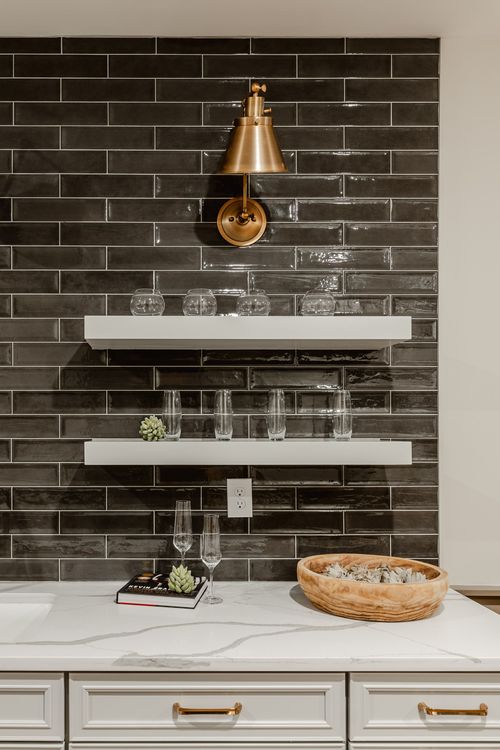 Wide view of a wet bar with black tile, triple gold sconces and floating glass shelves