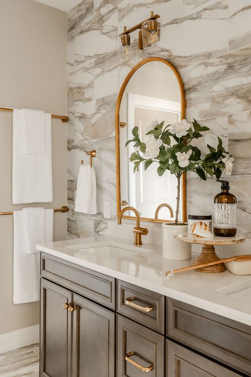 Primary bathroom vanity with marble tile wall, an arched brass mirror and fresh flowers