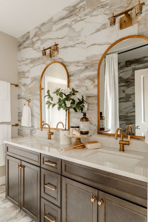 Double-sink bathroom vanity with marble tile wall and twin arched brass mirrors with gold sconces