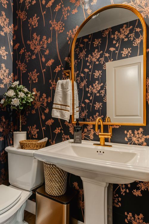 Powder room with dark floral wallpaper, an arched brass mirror and pedestal sink