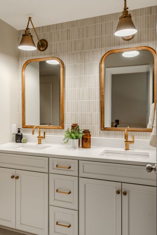 Double vanity bathroom with vertical tile wall, arched gold mirrors and gray cabinetry