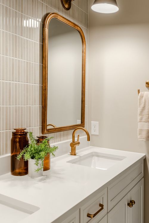 Powder room vanity with an arched gold mirror, vertical tile wall and amber glass jars with greenery