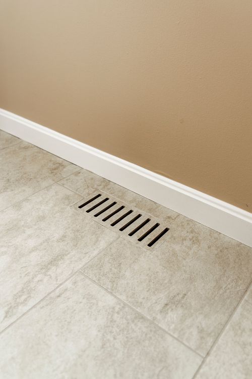 Close-up of a floor vent along a tan wall and stone-look tile floor