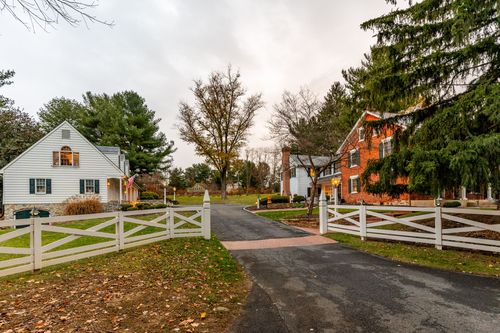 Driveway view of a white fenced entrance leading to a brick farmhouse and carriage house
