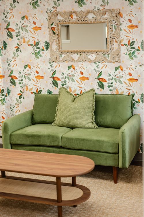 Close-up of a green loveseat with a fringed pillow against floral wallpaper
