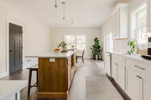 Long white kitchen with wood island and dark door leading toward a staircase and dining area