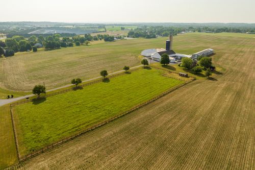 Aerial view of a farmstead bordered by a wood-fenced pasture and tree line, silo and barns beyond