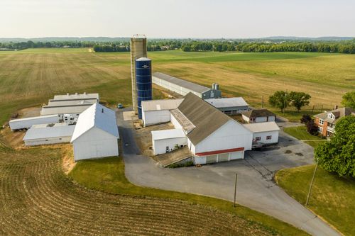 Aerial view of a farmstead with silo and barns beside a brick farmhouse and gravel drive