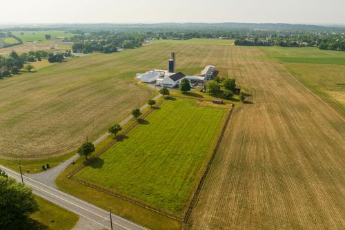Aerial view of a farm complex with silo and barns beside a fenced green pasture along a country road