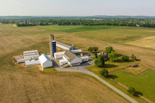 Aerial view of a farm complex with silo, barns and a distant brick farmhouse amid open fields