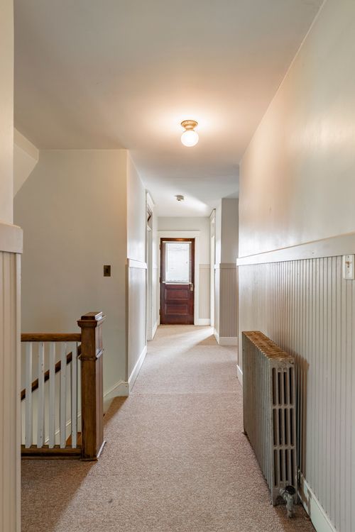 Hallway with a staircase newel post, wood door and radiator along a carpeted runner