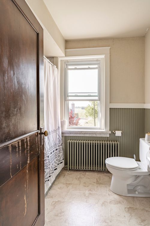 Bathroom with a dark wood door, patterned shower curtain, radiator and a window view of farmland