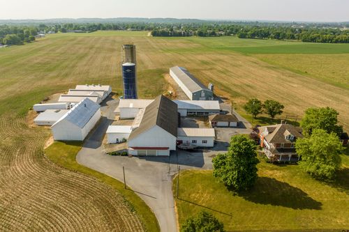 Aerial view of a farm complex with silo and connected barns beside a gravel driveway and fields