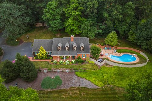 Aerial view of a brick country home with wraparound porch, pool, and wooded lot