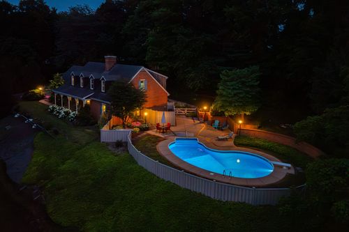 Aerial nighttime view of a lit home with pool beside a wooded backyard