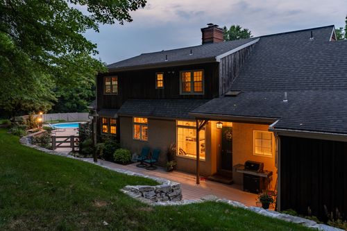 Aerial dusk view of a dark-shingled home with lit windows and a distant pool