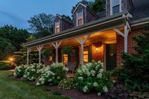 Twilight exterior of a home with dark wood dormers, brick, and hydrangea landscaping