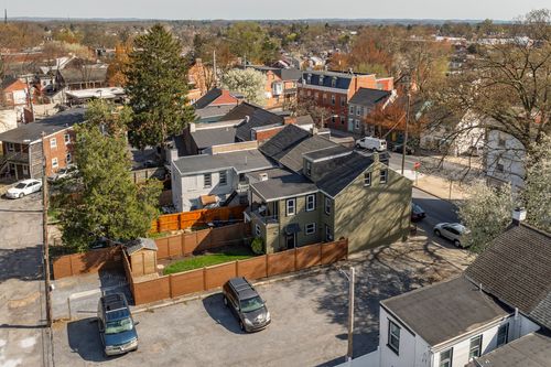 Aerial view of a corner property with fenced yard, parked cars, and city rooftops beyond