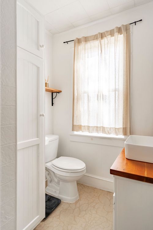 White bathroom with a vessel sink on a wood countertop, sheer tan curtains and beadboard walls.