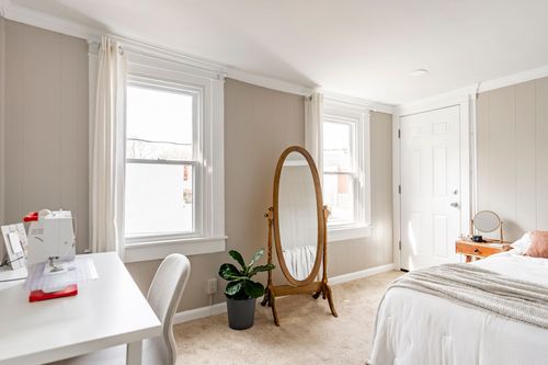 Bedroom set up as a sewing room with a craft desk, oval floor mirror and two bright windows.
