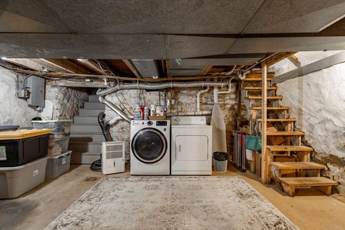 Unfinished stone-walled basement with a washer and dryer beneath open joists and wood stairs.