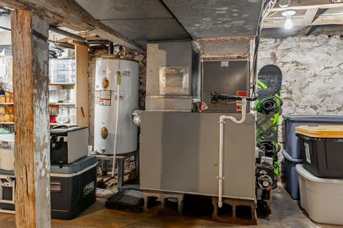 Basement utility area with a water heater, ductwork and gear stored along stone walls.