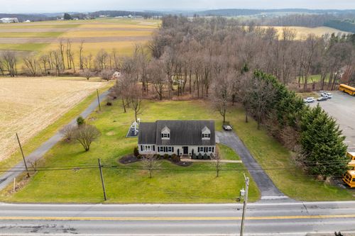 Aerial view of a cape-style home with a paved driveway, mature trees and farmland beyond.