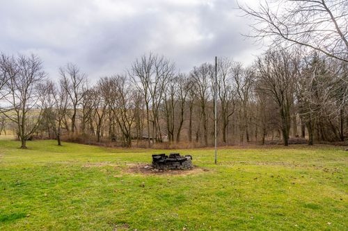 Backyard lawn with a stone fire pit, flagpole and a wooded tree line beyond.