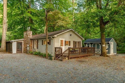 Cabin exterior with a stone chimney, tan siding and a covered deck surrounded by mature woods.