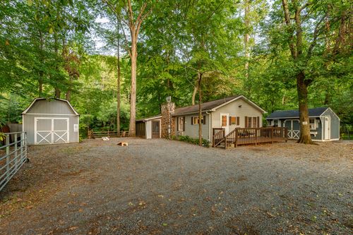 Cabin exterior with a stone chimney, storage sheds and a wooded gravel driveway.