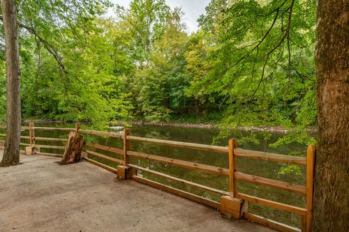 Creekside path with a wood rail fence and tree stump, framed by green woods.