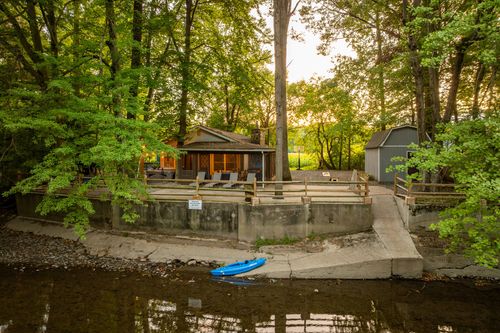 Riverside view of a cabin and matching storage shed nestled among trees at sunset, kayak on the bank.