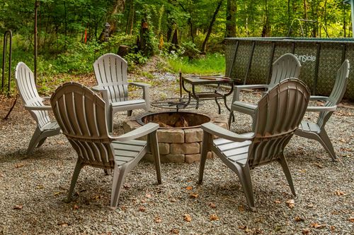 Circle of adirondack chairs around a lit stone fire pit in a gravel clearing surrounded by trees.